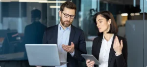 Two business professionals discussing strategies while standing in front of a laptop in a modern office setting