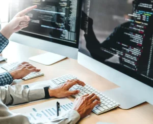 Two individuals collaborating at desks, focused on their computers displaying lines of code