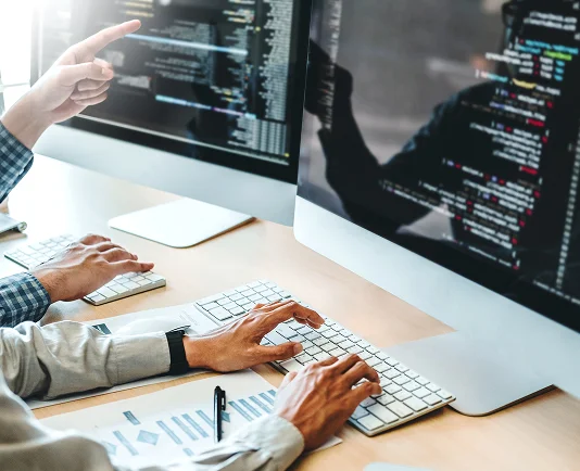 Two individuals collaborating at desks, focused on their computers displaying lines of code